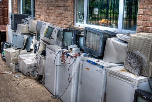 A row of old, discarded electronic devices, primarily various models of CRT computer monitors and desktop towers, is lined up against a brick building exterior with large windows reflecting greenery. The monitors display different screen images, and some have visible wires and power cables hanging loosely. The equipment shows signs of wear, with some screens cracked or cloudy, and the casings are made of grey or beige plastic with textured finishes. The collection is placed directly on the pavement outside a building, indicating a disposal or collection site for waste electronic equipment. In the background, the brick wall has a window with a white frame and a partially visible interior. The scene exemplifies electronic waste that generic rubbish removal services, such as those provided by Office Clearance Ealing, may handle as part of workplace clearance efforts.