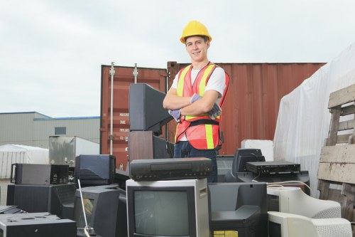 A young male worker dressed in a white t-shirt, high-visibility safety vest with yellow and orange stripes, and a yellow safety helmet, standing outdoors at an industrial site or storage yard. Behind him, there is a large brown shipping container, and to the right, a stack of wooden pallets or crates. In the foreground, numerous discarded electronic waste items are visible, including old computer monitors, keyboards, and other obsolete electronic devices, all positioned on the ground and some stacked beneath each other. The environment appears clear and bright with overcast sky, indicative of a waste clearance operation typical of a rubbish removal service in the Ealing area, aimed at clearing office or workplace clutter. The individual is smiling, suggesting a professional and organized approach to rubbish removal, with the focus on electrical and office equipment disposal, aligning with services offered by companies like Office Clearance Ealing.