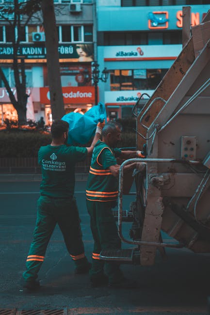An outdoor scene on a tree-lined street showing a waste collection vehicle with an open rear compartment, which appears weathered and has residual debris inside. A worker dressed in a green uniform wi