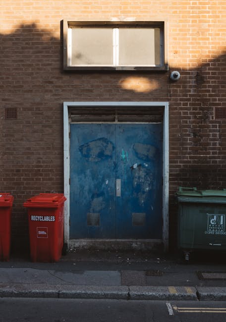 A weathered blue metal door with visible scratches and patches of paint peeling, set into a brick wall with reddish-brown bricks. Above the door, a small rectangular window with frosted glass is frame
